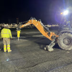 Two workers in high-visibility clothing observe a backhoe operating at night on an illuminated worksite.