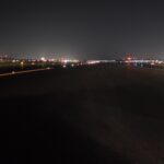 A dark airport runway at night with distant city lights and illuminated runway markers visible.