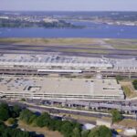 Aerial view of an airport terminal with parked airplanes, runways, a parking garage, and a river in the background.