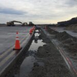 Construction site on a paved road with orange cones, machinery, and a trench filled with water and dirt.