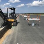 A construction vehicle works on a paved road with traffic cones and barriers set up under a partly cloudy sky.
