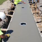 Construction workers smoothing freshly poured concrete on a sidewalk with circular utility access points.
