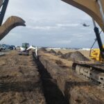 Excavators digging a long trench in a field with a helicopter flying in the cloudy sky above.