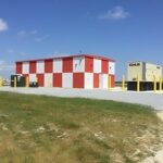 A red-and-white checker-patterned building stands next to a beige generator in a fenced, grassy area under a blue sky.