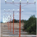 A row of orange airport approach lights on metal poles, lined up along a dirt path under a cloudy sky.