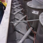 Several metal pipes and cylinders are connected with rods inside a rectangular metal frame; a worker stands nearby.