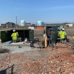 Two utility workers in safety gear work on open electrical panels at a construction site near a fenced area.