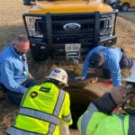 Four workers kneel and observe an open manhole in front of a yellow utility truck on grass.