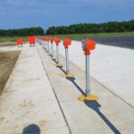 Row of orange runway approach lights mounted on poles along a concrete surface near a grassy area and a parked vehicle.