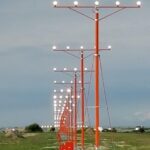 A row of red approach lights on tall poles lines a grassy area near an airport runway under a cloudy sky.
