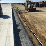 Construction workers and machinery work on a road edge, installing drainage next to a newly paved concrete surface.