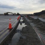 Construction site with orange traffic cones, excavation, muddy water, and heavy machinery under a cloudy sky.