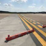 Concrete and asphalt airport taxiway with yellow lines and red barricades under a partly cloudy sky.