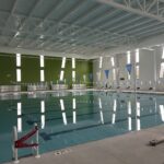 Indoor swimming pool with lane dividers, starting blocks, and a green accent wall; poolside area is empty.