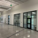 Large empty hallway with polished concrete floors, high ceiling, and a row of offices with black-framed glass windows and doors.