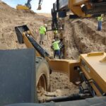 Construction workers in safety vests operate heavy machinery while working in a trench at a sandy construction site.