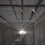 Concrete tunnel with metal conduit pipes attached to the ceiling and light visible at the far end of the tunnel.
