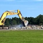 A yellow excavator moves rocks on a construction site with trees and an American flag in the background.