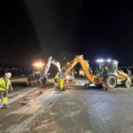 Workers and two excavators perform nighttime maintenance on an airport runway under bright floodlights.