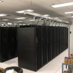 Rows of black server racks in a data center room with overhead cable management and supplies visible in the foreground.