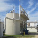 Rectangular and cylindrical tanks with white pipes at a water treatment facility under a partly cloudy sky.