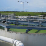 A wastewater treatment facility with large circular tanks, metal walkways, and pipes, set against a grassy landscape.