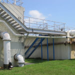Outdoor view of a water treatment facility with large pipes, metal stairs, and a grassy area under a clear sky.