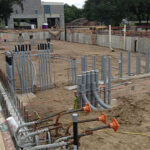 Construction site with exposed conduit pipes, rebar, and foundation work in progress for a new building.