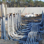 Rows of gray electrical conduit pipes are installed underground at a construction site with equipment in the background.